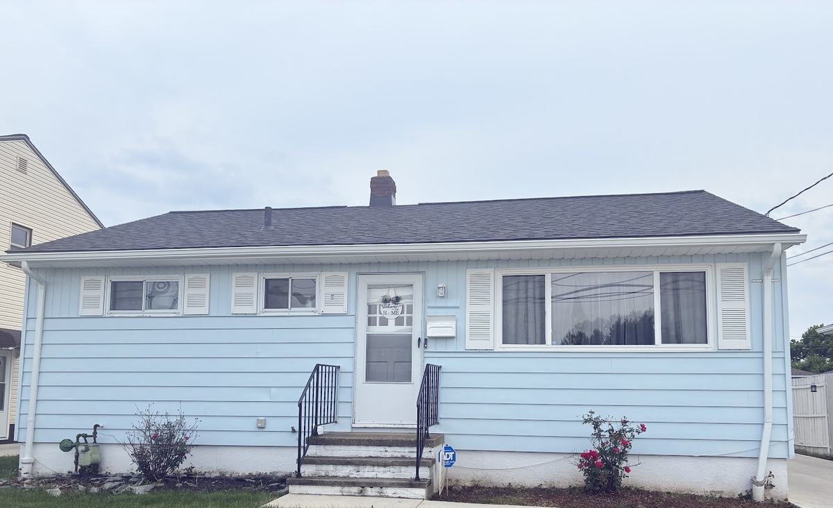 A ranch house with pale blue siding, white shutters and trim, under a gray sky.