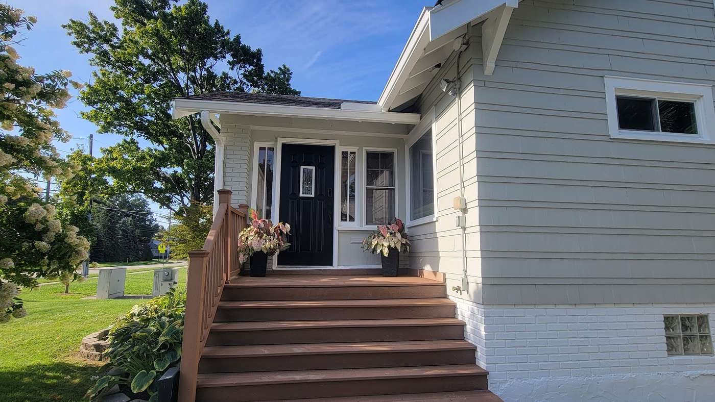 A gray-sided house with a black front door, white trim and brown wooden steps, with planters of colorful leaves by the door.