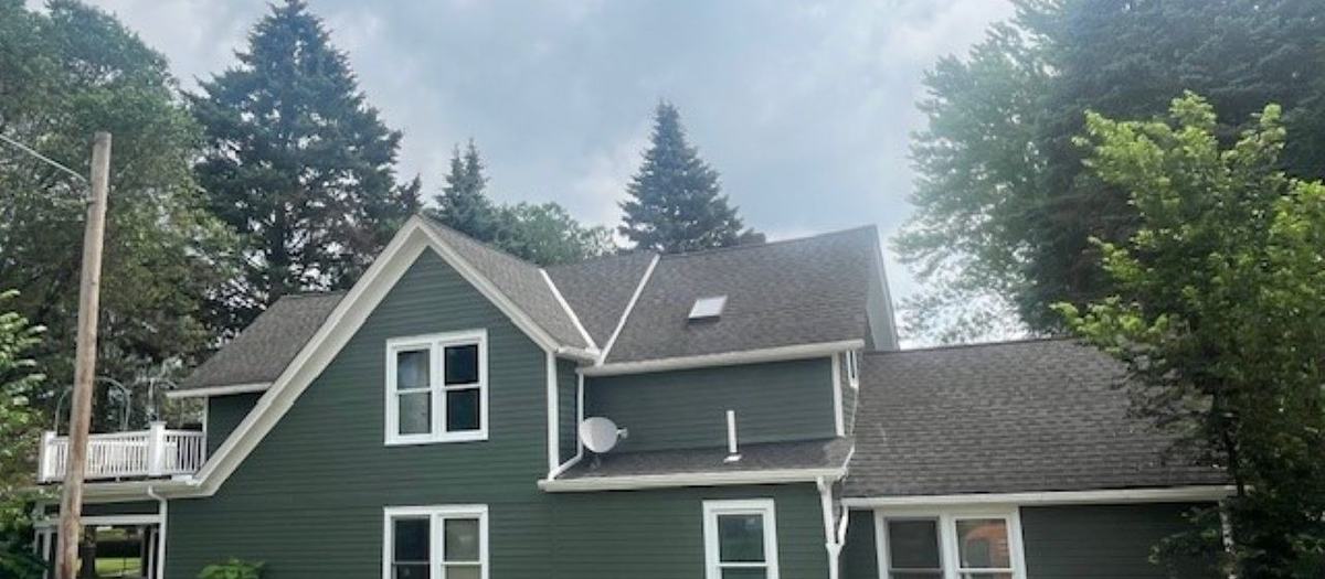 A two-story house with green siding and white trim, with tall evergreen trees behind it.